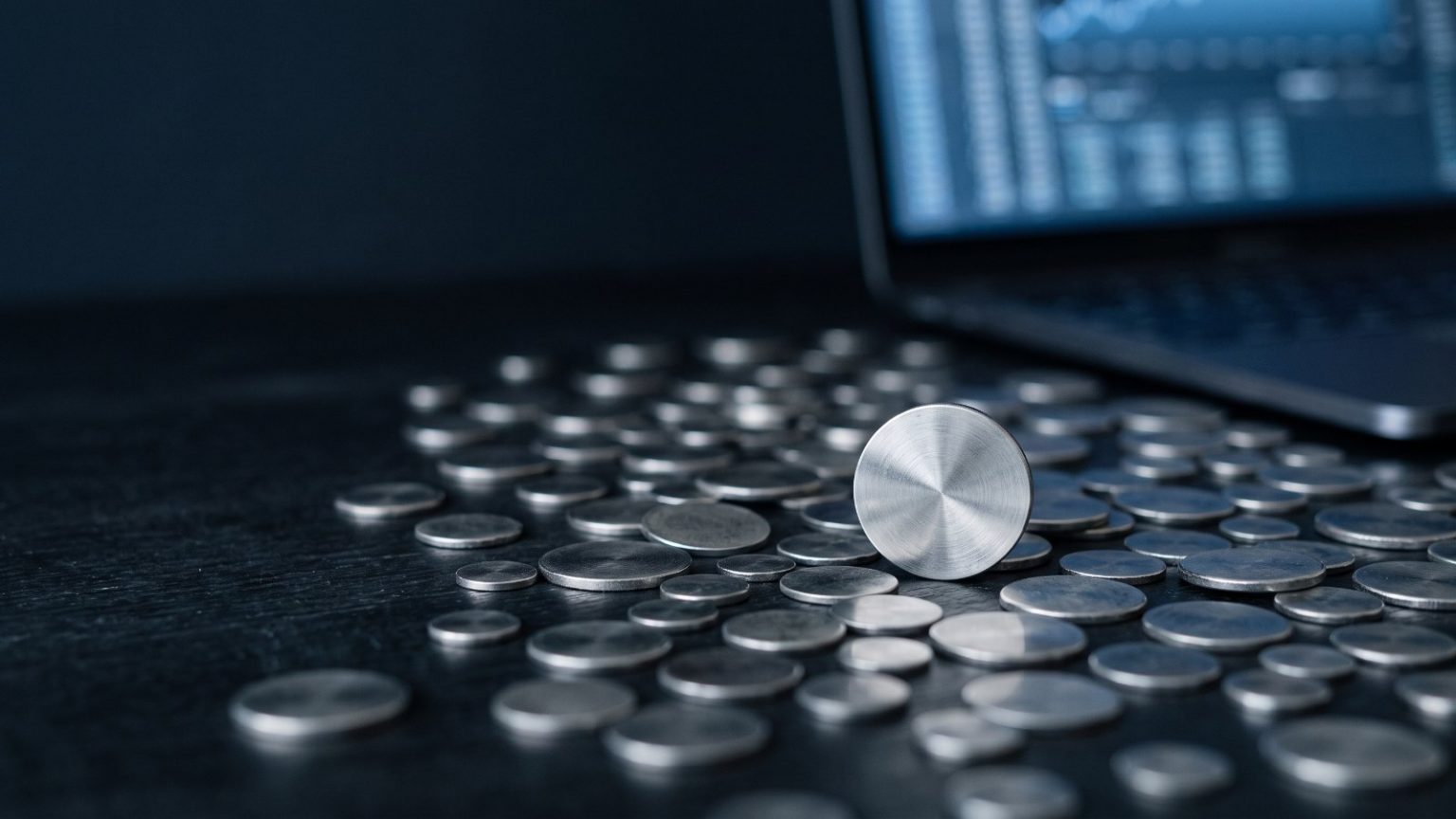 Diversas moedas metálicas espalhadas sobre mesa escura com notebook ao fundo, representando as altcoins